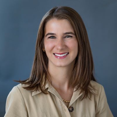 Smiling light-skinned, female-presenting individual with medium length brown hair wearing a tan shirt on a grey backdrop.