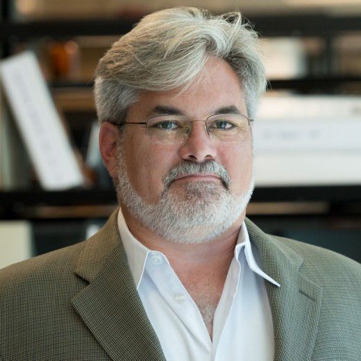 A light-skinned male-presenting individual with gray hair and a beard wearing a gray suit jacket and white button-up shirt against a blurred backdrop