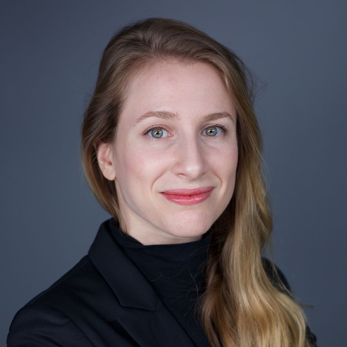 Smiling light-skinned, female-presenting individual with long length blonde hair wearing a black shirt on a grey backdrop.