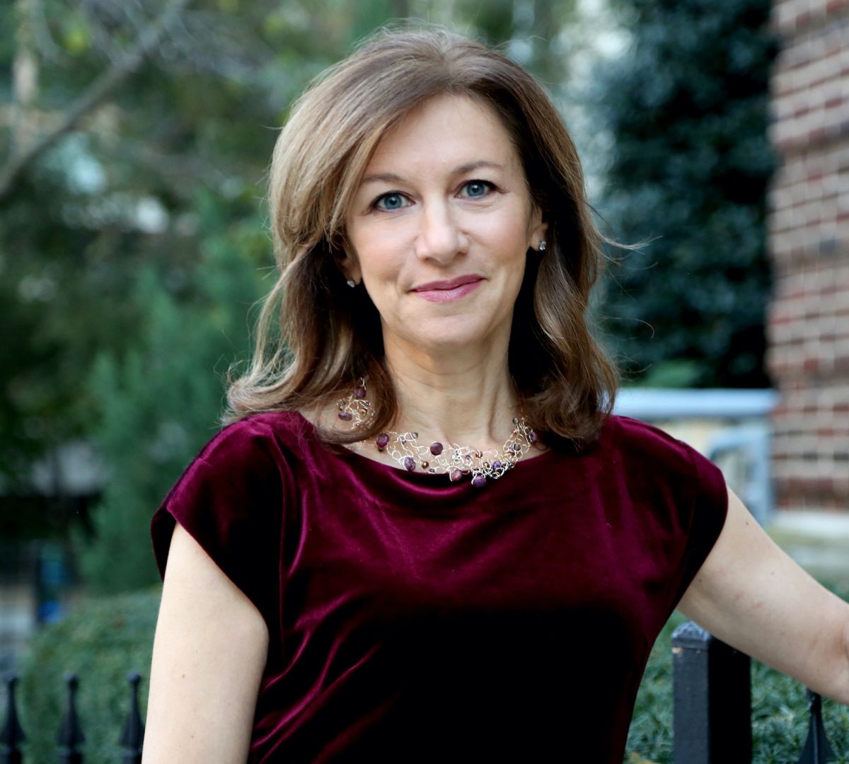 Smiling medium-skinned, female-presenting individual with medium length light hair wearing a burgundy shirt in an outdoor setting