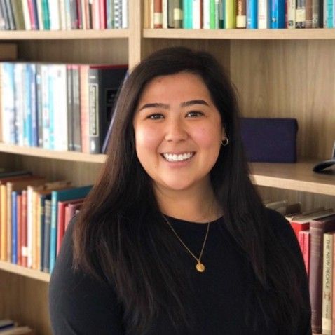 A smiling light-skinned female-presenting person with dark brown hair wearing a black shirt with a bookshelf in the background