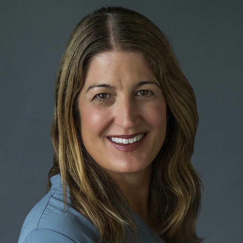 Smiling light-skinned, female-presenting individual with medium length dark hair wearing a blue shirt on a grey backdrop.