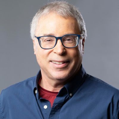 Smiling light-skinned, male-presenting individual with short grey hair wearing a blue shirt on a grey backdrop.