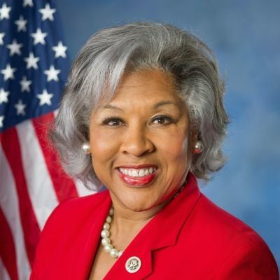 Smiling dark-skinned, female-presenting individual with medium length grey hair wearing a red suit, American flag and blue background