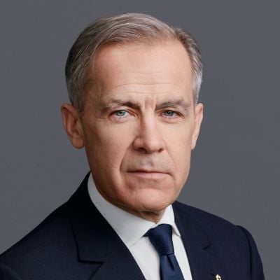 Smiling light-skinned, male-presenting individual with short grey hair wearing a dark suit on a grey backdrop.