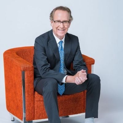 Smiling light-skinned, male-presenting individual with short grey hair wearing a dark suit seated in an orange chair.