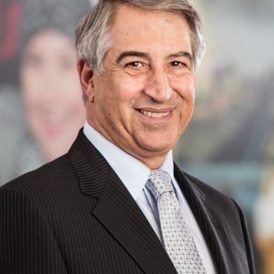 Smiling light-skinned, male-presenting individual with short grey hair wearing a dark suit on a grey backdrop.