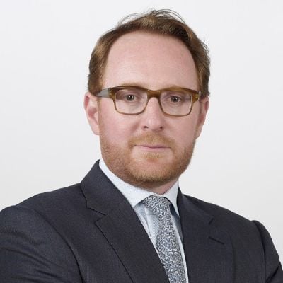 Smiling light-skinned, male-presenting individual with short red hair wearing a dark suit on a white backdrop.