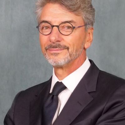 A smiling light-skinned male with gray hair and a beard wearing a dark suit with glasses against a gray background