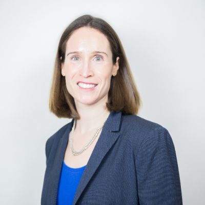 Smiling light-skinned, female-presenting individual with medium length light brown hair wearing a blue suit on a white backdrop.