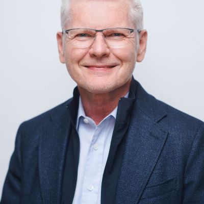 Smiling light-skinned, male-presenting individual with short grey hair wearing a dark suit on a grey backdrop.