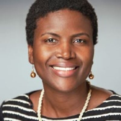 Smiling darl-skinned, female-presenting individual with short dark hair wearing a black and white striped shirt on a grey backdrop.