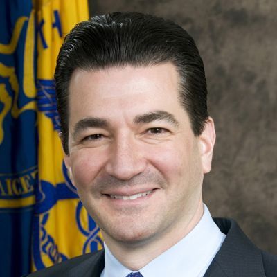 Smiling light-skinned, male-presenting individual with short dark hair wearing a dark suit in front of a flag