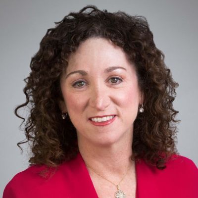 A smiling light-skinned female with dark brown curly hair wearing a red shirt against a light gray background