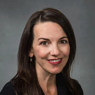 A smiling female-presenting light-skinned person with dark brown hair wearing a dark shirt against a gray gradient background