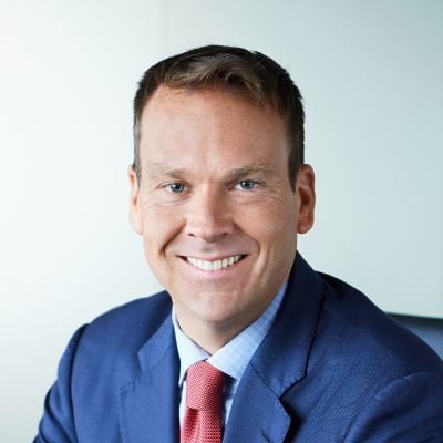 Smiling light-skinned, male-presenting individual with short dark hair wearing a blue suit on a grey backdrop.