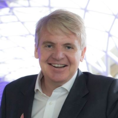 A smiling light-skinned male-presenting person with white hair wearing a dark suit jacket against a blurred background