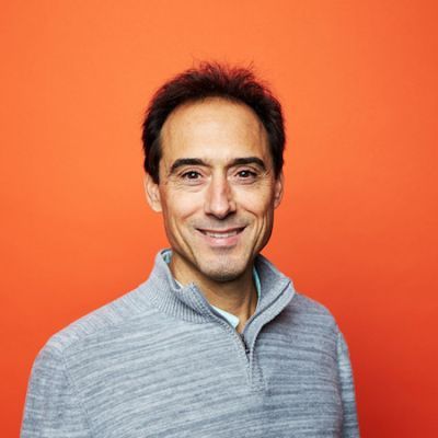 A smiling light-skinne male with dark hair wearing a gray sweater against a bright orange background