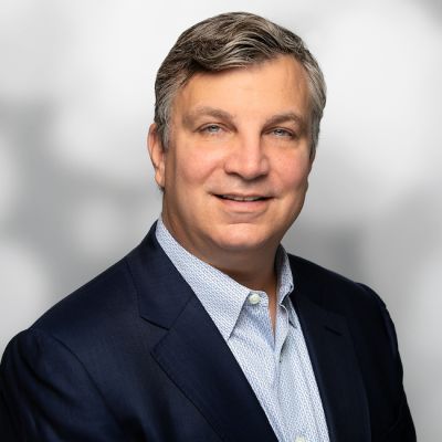 Smiling light-skinned, male-presenting individual with short grey hair wearing a dark suit on a grey backdrop.