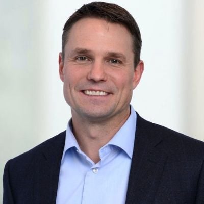 Smiling light-skinned, male-presenting individual with short dark hair wearing a dark suit on a grey backdrop.