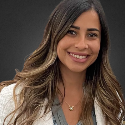 Smiling female-presenting individual with long dark hair wearing a white jacket on a grey backdrop.