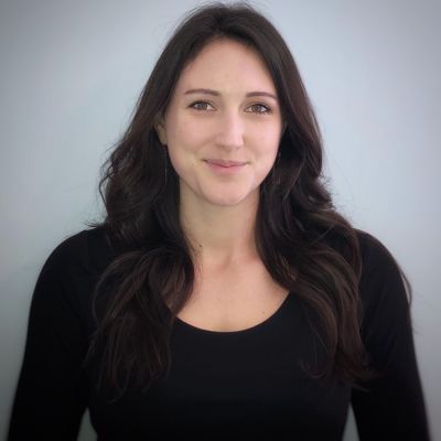 Smiling light-skinned, female-presenting individual with long dark hair wearing a black shirt on a grey backdrop.