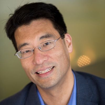 A smiling male of Asian heritage wearing glasses and a navy suit