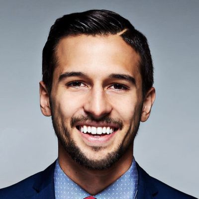 A smiling lighter-skinned male-presenting person with dark brown hair and a beard wearing a suit against a light gray background