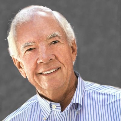 A smiling light-skinned male with white hair wearing a blue stripe button-up against a dark gray background