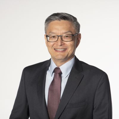 Smiling male-presenting individual with short grey hair wearing a dark suit on a grey backdrop