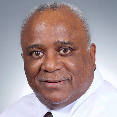 A smiling dark-skinned male-presenting individual with gray hair wearing a white buttonup against a gradient background