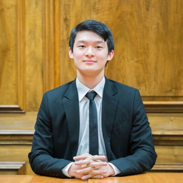 A smiling male-presenting person of Asian heritage with black hair wearing a black suit and tie against a wooden background