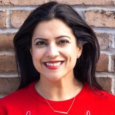 A smiling female-presenting person with dark brown hair wearing a red shirt against a brick background