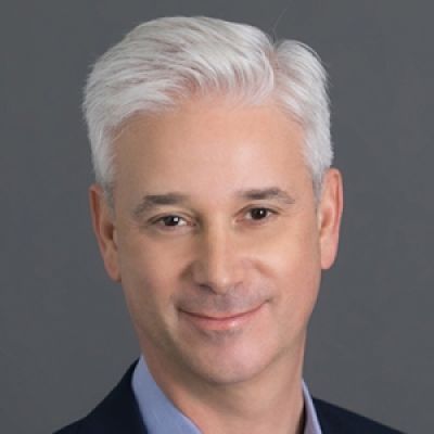 Smiling light-skinned, male-presenting individual with short grey hair wearing a dark suit on a white backdrop.