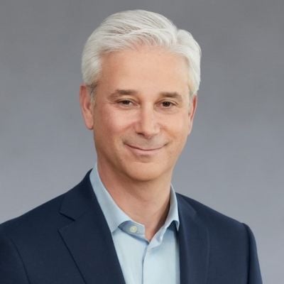 A smiling light-skinned male with white hair wearing a navy suit jacket over a light blue buttonup against a gray gradient background