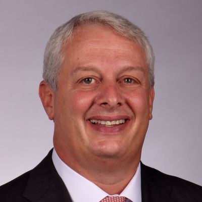 A smiling light-skinned male with gray hair wearing a dark suit against a light gray background