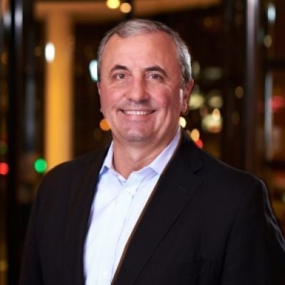 Smiling light-skinned, male-presenting individual with short grey hair wearing a dark suit on an indoor background