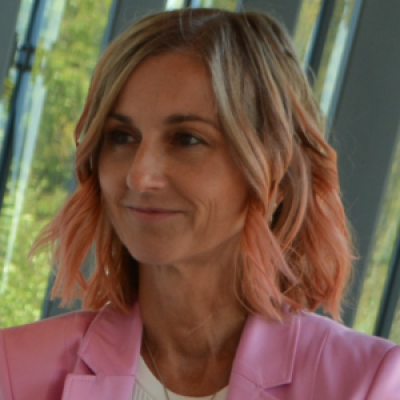 A light-skinned woman with blonde hair that has pink at the tips wearing a pink suit jacket against an outdoor background