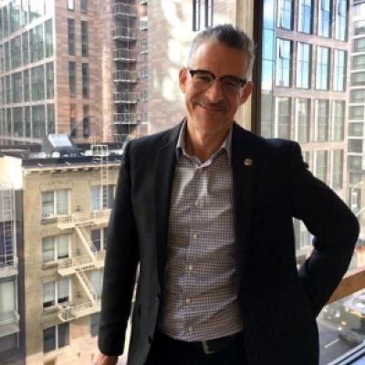 A smiling light-skinned male with gray hair wearing glasses and a dark suit jacket over a button-up shirt against a city skyline backdrop