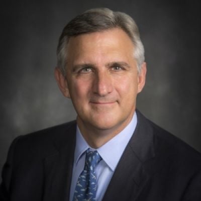 A smiling light-skinned male with gray hair wearing a dark suit and tie against a dark gray background