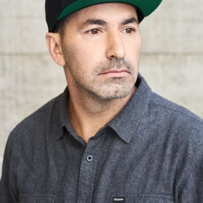 A light-skinned male wearing a dark gray button-up and a flat-billed hat against a blurred background