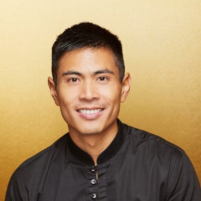 A smiling male of Asian heritage wearing a black shirt against a gold background