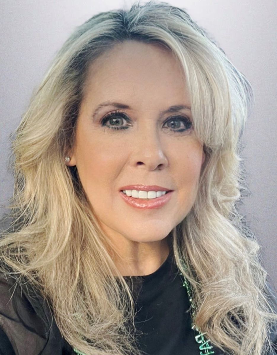 Smiling light-skinned, female-presenting individual with blonde hair wearing a dark shirt on a grey backdrop.