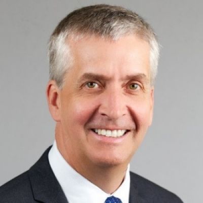 A smiling light-skinned male with white and gray hair wearing a dark suit and tie against a gray background