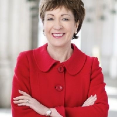 A smiling light-skinned female with dark brown hair wearing a red suit jacket against an ambiguous background