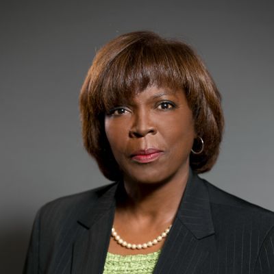 A darker-skinned female with short brown hair wearing a black suit jacket over a green shirt and pearls against a gray background