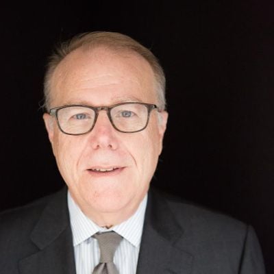 A light-skinned male wearing glasses and a dark gray suit and tie against a black backdrop