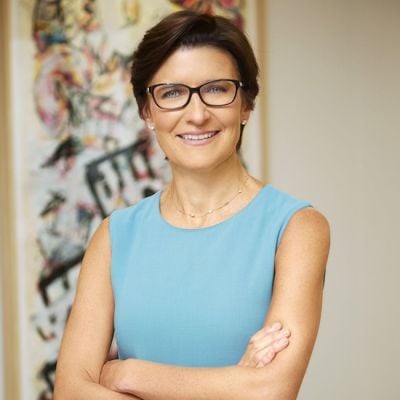 A smiling light-skinned female with dark brown hair wearing glasses and a blue shirt against an abstract background