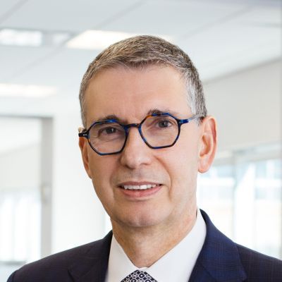 A smiling light-skinned male with gray hair wearing glasses and a navy blue suit against an ambiguous background