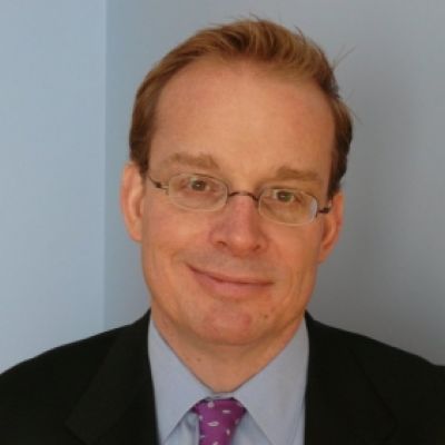 A light-skinned male with brown hair wearing glasses and a dark suit and tie against a light blue background